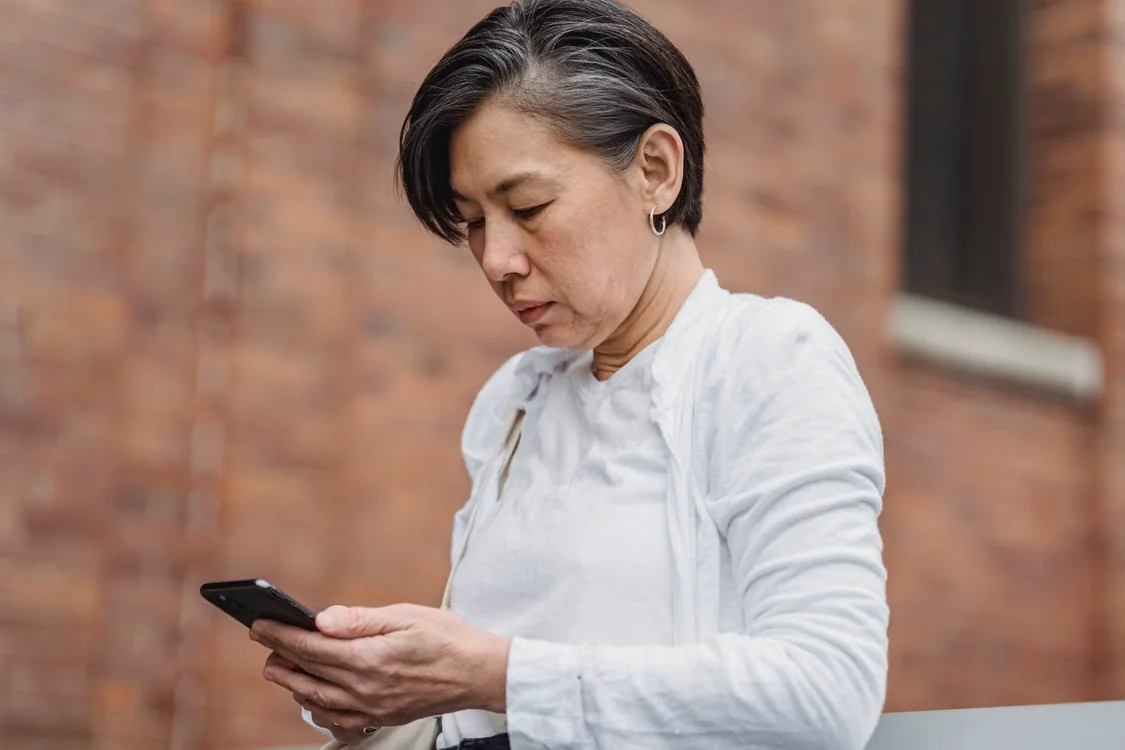 A middle-aged woman intently uses her smartphone against a brick wall backdrop.