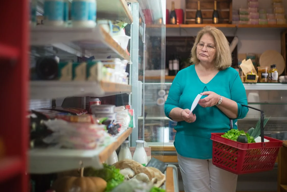 Older woman shopping with basket and list in grocery store aisle.