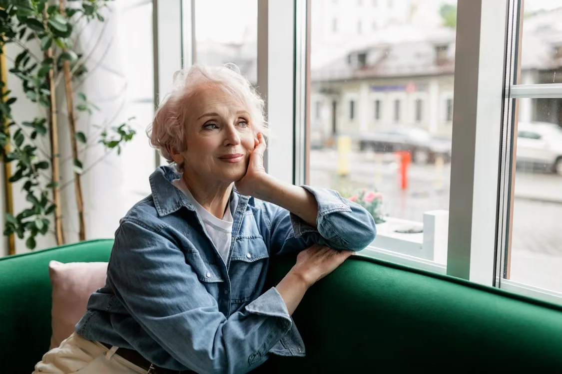 Graceful senior woman in denim jacket sitting by a window, reflecting quietly indoors.