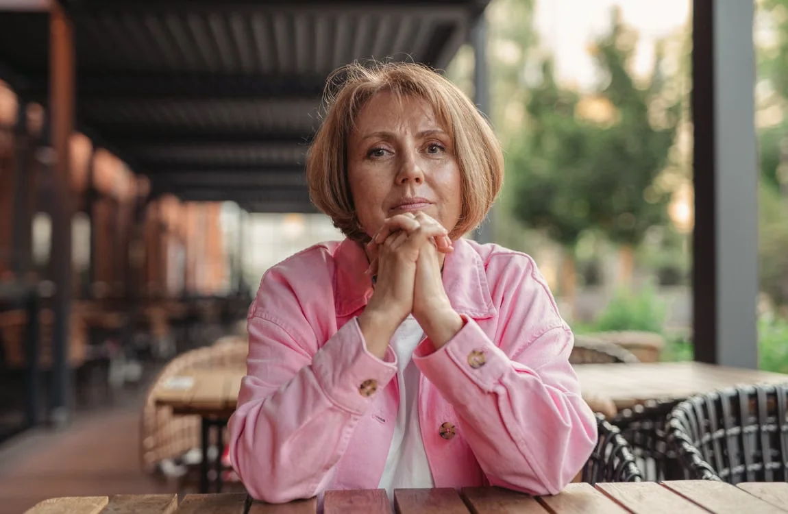 Portrait of a senior woman in pink, sitting outdoors in a contemplative pose.