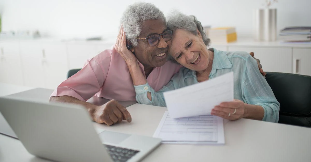 A joyful senior couple smiling while reviewing retirement income documents together at a desk with a laptop