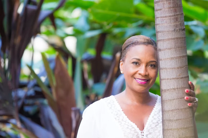 A smiling black middle-aged woman in a white lace shirt leaning against a tree