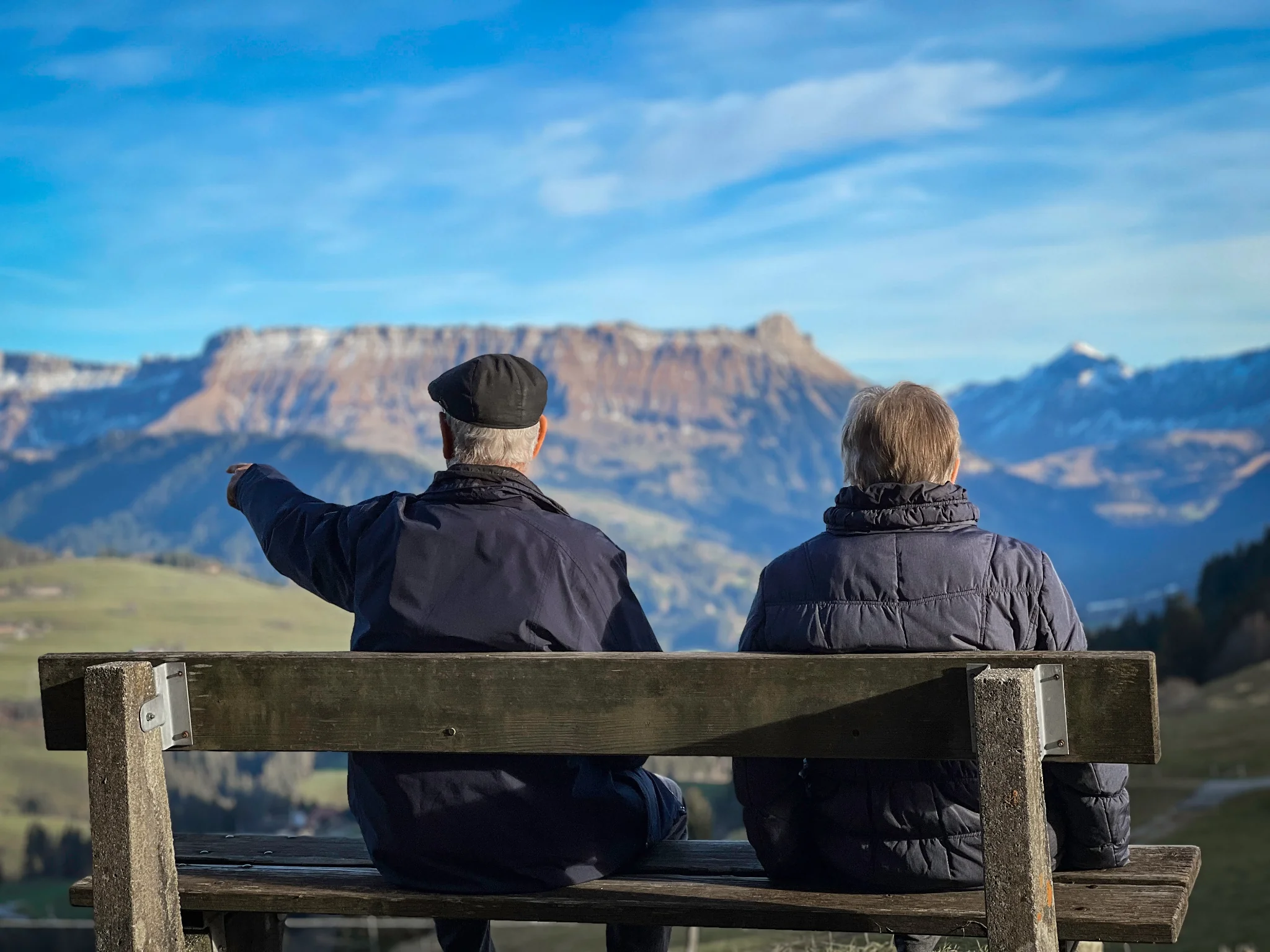 Senior couple sits on bench admiring a mountainous landscape.