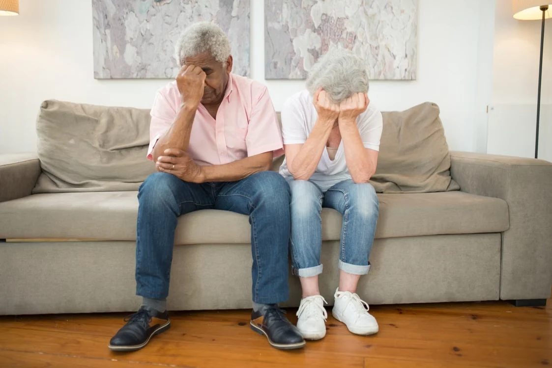Elderly couple sitting on a sofa, annoyed with their heads in their hands.